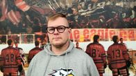 En man med glasögon och grå huvtröja står vid en ishockeyrink med personer i ishockeykläder bakom sig på isen.