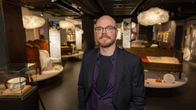 Kuvassa henkilö poseeraa museon näyttelytilassa, jonka taustalla on esillä erilaisia huonekaluja ja esineitä.