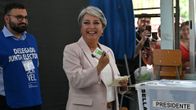 Jakkupukuun pukeutunut hymyilevä nainen pitelee käsissään äänestyslipukkeita äänestyspaikalla.