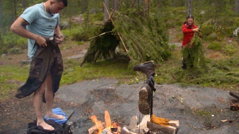 Mies laittaa housuja jalkaan ja nainen tekee oksista telttaa metsässä.