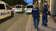 Kuvassa näkyy kaksi poliisia seisomassa kadulla, ja taustalla on useita poliisiautoja, jotka ovat parkkeerattu tien varteen. 