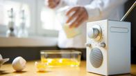 Keittiönpöydällä etualalla radiovastaanotin, jonka vieressä kananmunankuoria. Ihmishahmo mittaa taustalla sokeria, aikoo leipoa jotain.