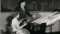 Autokoulun opettaja katsoo karttakeppi kädessään, kun nainen ajaa vanhanaikaisella (vuonna 1957) ajosimulaattorilla.
