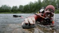 Laitesukeltaja asettaa kivelle jokihlemisimpukan. Sukeltaja makaa matalassa vedessä. Yllä punainen sukelluspuku.