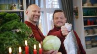 Kuvassa kaksi henkilöä pitelee kädessään kaalia ja kalaa. Etualalla on joulukynttelikkö. Taustalla näkyy keittiö, jossa on hyllyllä astioita.