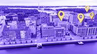 Violetissa kaupunkimaisemassa näkyy kerrostaloja veden ympäröimänä. Neljän kerrostalon yläpuolella leijuu keltainen sijaintitäppä.