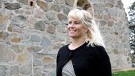 Nainen hymyilee ja katsoo kuvasta vasemmalle. Taustalla vanhan kirkon kiviseinää.