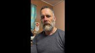 A tried, sad-looking middle-aged man with a grey beard and T-shirt looks into the camera in a ship cabin. 