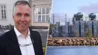 En man i kostym står utomhus framför en byggnad; till höger syns en industribyggnad vid vatten.