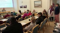 Useita ihmisiä istuu valtuustosalissa seuraamassa esitystä, jonka taululla näkyy tekstiä 10.09.2026 Utsjoella..