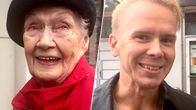 Iäkäs nainen punaisessa takissa ja mustassa baskerissa sekä nuori mies mustassa takissa seisovat ulkona rakennuksen edessä.