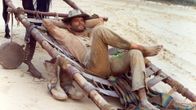 Hymyilevä lännenasuinen mies (näyttelijä Terence Hill) makailee hevosen valjaisiin kiinnitetyllä kenttävuoteella tai paarilla jalka toisen polven päällä, kädet ristissä pään takana ja hattu silmillä. Kuva elokuvasta Trinity ratsastaa jälleen.