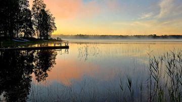 Tyyni järvi, jonka rannalla on laituri ja veneitä, taustalla metsää sekä aamuruskon värejä taivaalla.