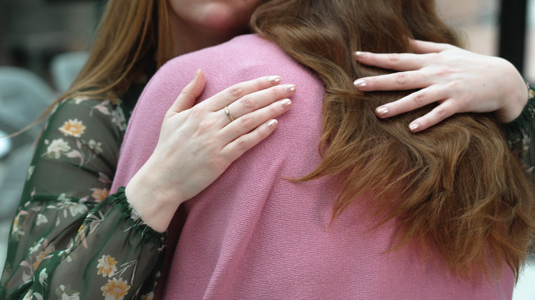 Two women hugging, faces are not visible. One is in a pink jumper and the other is wearing a flowered green shirt.