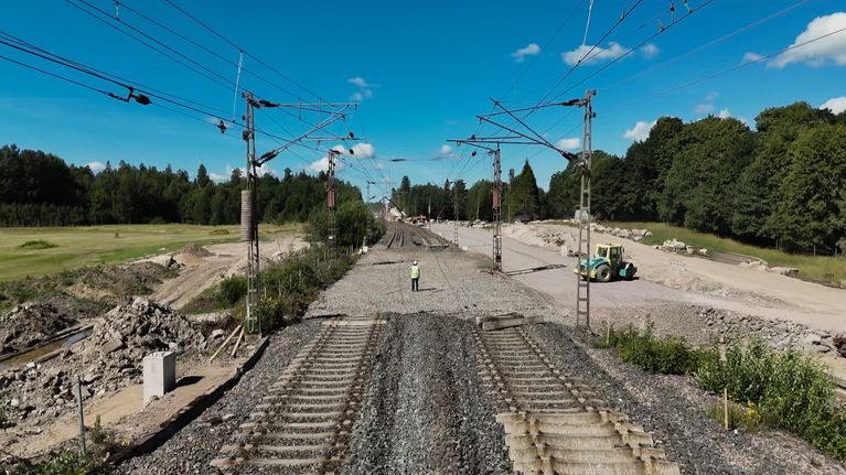 A railway construction site with two sections of incomplete track, a worker and a tractor with fields and trees on either side.