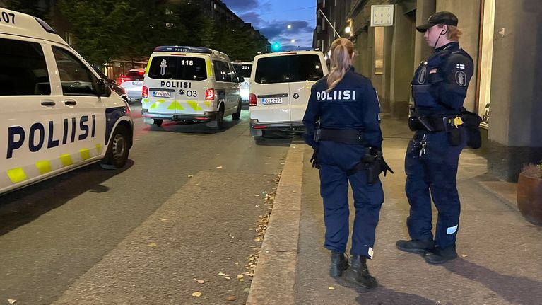 Kuvassa näkyy kaksi poliisia seisomassa kadulla, ja taustalla on useita poliisiautoja, jotka ovat parkkeerattu tien varteen. 