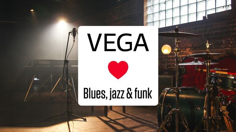 En vit skylt där det står Vega ❤️ blues, jazz och funk, i bakgrunden finns ett trumset och en mikrofon på stativ.
