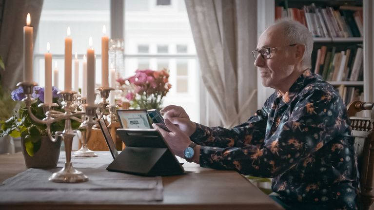 Harmaapäinen seniorimies näpyttelee ruokapöydän ääressä älypuhelintaan. Pöydällä on myös tabletti ja läppäri.