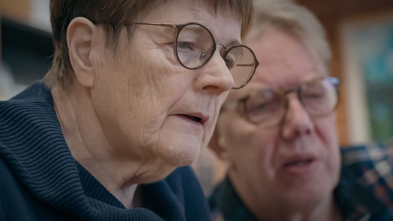 Etualalla lähikuvassa silmälasipainen seniorinainen. Taustalla epätarkkana harmaapäinen seniorituutorimies.