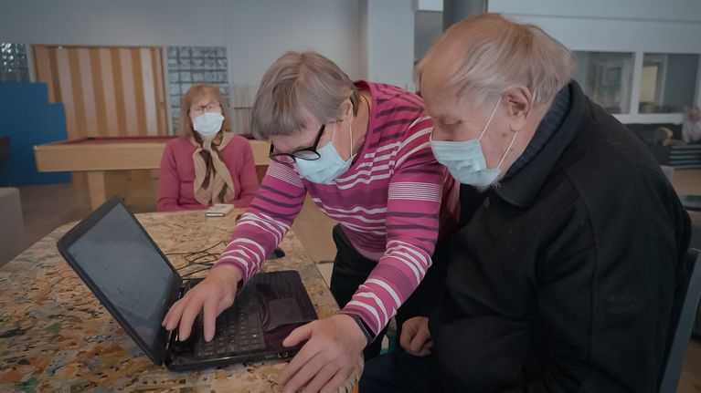 Seniorimies istuu läppärin edessä pöydässä. Läppärille on sivusta kumartunut nainen, joka näppäilee näppäimistöä. Taustalla kauemmalla näkyy toinen nainen istumassa pöydän päässä.