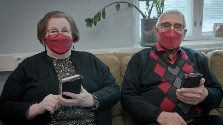 Seniorinainen ja seniorimies istuvat sohvalla puolikuvassa älypuhelimet käsissään ja katsovat kameraan.