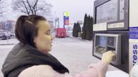 Nainen laittaa pankkikorttia bensiiniautomaatin lukijaan, taustalla luminen maisema aamuhämärässä ja huoltoaseman hintataulu.