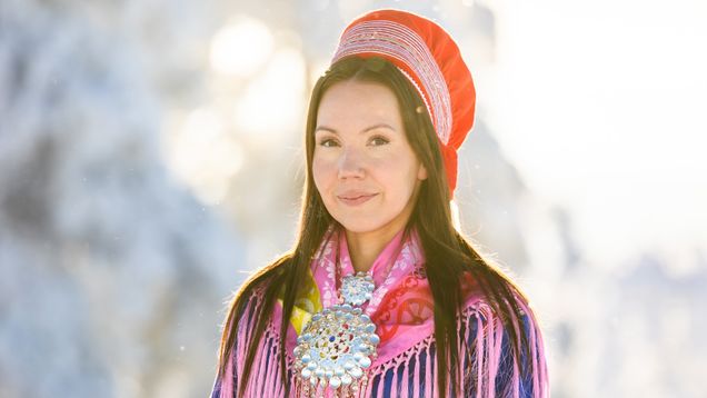 Anne-Maria Magga seisoo talvisessa maisemassa, yllään perinteinen saamelaisasu ja suuri hopeakoriste rinnassaan.
