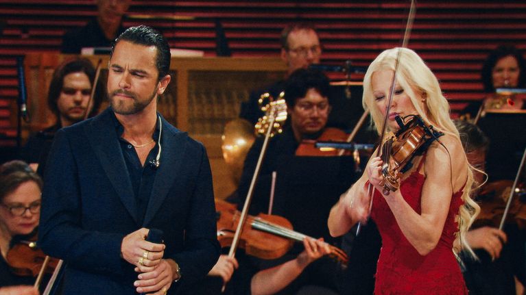 Laulaja ja viulisti esiintyvät orkesterin säestyksellä konserttilavalla.