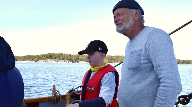 Tom Westerlund står vid rodret tillsmamans med en skolelev. Soligt försommarväder.