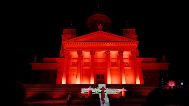 Näyttelijöitä seisoo Helsingin tuomiokirkon rappusilla, johon on heijastettu ristin muodossa oleva valokeila. Kirkko on valaistu punaisilla valoilla.