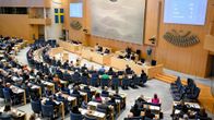 Riksdagens kammare med ledamöter sittande i bänkar, på väggen syns en svensk flagga och en skärm med siffror och text.