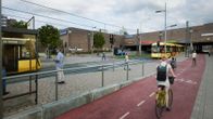 Personer går och cyklar på en livlig stadsplats med spårvagnar i bakgrunden.