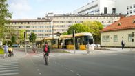 Personer går och cyklar på en livlig stadsplats med spårvagnar i bakgrunden.