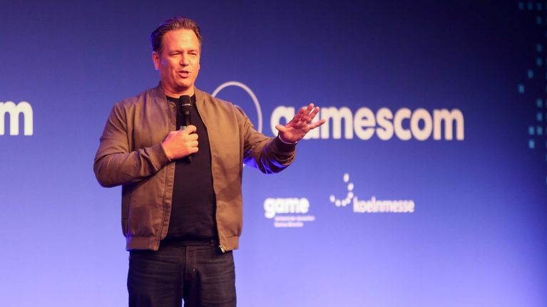 Phil Spencer puhuu mikrofoniin Gamescom-tapahtumassa, taustalla sininen mainosseinä.