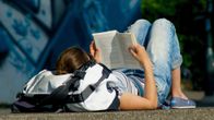 En person i jeans och randig tröja ligger på rygg utomhus och läser en bok, med en ryggsäck bredvid sig.