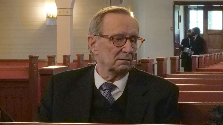 Mauri Päivinen istuu kirkon penkillä puku yllään ja katsoo viistosti alaspäin. Taustalla kaksi miestä kirkon sisäänkäynnin edessä.