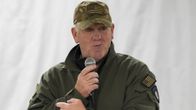Tom Homan puhuu mikrofoniin. Hänellä on yllään Yhdysvaltain lipulla koristettu maastokuvioinen lippalakki ja vihreä pilottitakki.