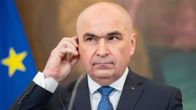 Tummaan pukuun pukeutunut kalju mies pitelee kuuloketta korvallaan. Taustalla näkyy EU:n lipun reuna.