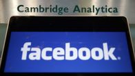 Facebookin logo näkyy näytöllä. Taustalla on kyltti, jossa lukee Cambridge Analytica.