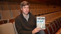 Mies istuu auditoriassa ja pitelee kirjaa pystyasennossa käsissään, näyttäen kantta kameralle.