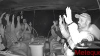 Black and white video capture of about a dozen people wearing life vests seated on a boat and holding their hands in the air.