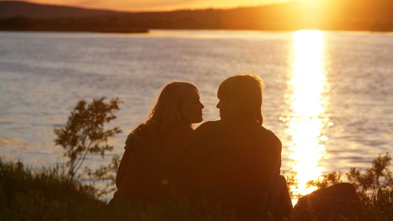 Silhouette of a couple sitting by a lake with golden sun shining on the horizon and the water.