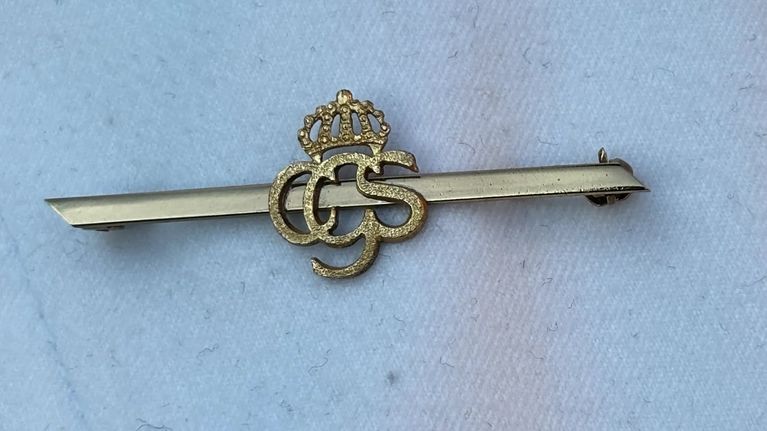 En brosch med krona och bokstäverna "CGS" är fäst på en ljus jacka bredvid ett pärlhalsband.