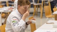 Kokki maistaa lusikalla uutetta pienestä valkoisesta astiasta pöydän ääressä.