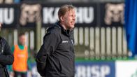 En fotbollstränarre i svart jacka ser ut över planen.