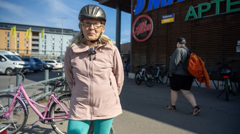 En äldre kvinna i rosa jacka och cykelhjälm står utanför en affär.