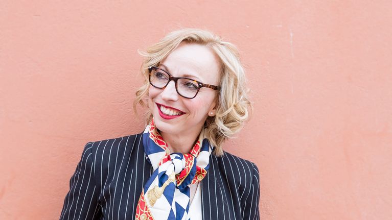 Maria-Elena Ehrnrooth, en dam med glasögon och ljust, lockigt hår står vid en rosa vägg.