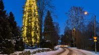 Valaistu, iso joulukuusi ulkona lumisessa kylämaisemassa. Tie kiemurtelee kuusen oikealla puolella. 