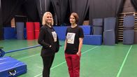 Kaksi ihmistä seisoo sisätiloissa liikuntatiloissa käymässä keskustelua. Heillä on päällään printtipaidat, joissa lukee Taidekoulu Estradi.