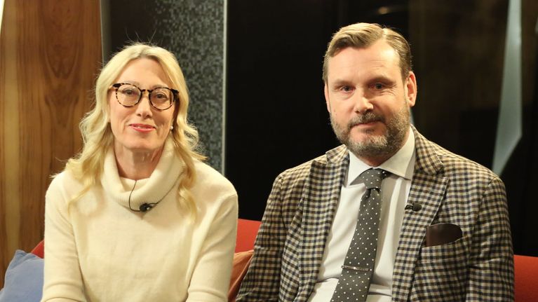 Kuvassa nainen ja mies istuvat punaisella sohvalla ja katsovat kameraan. Naisella on päällään vaalea villapusero, miehellä kauluspaita ja kravatti.